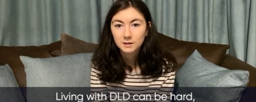 Video screenshot with a girl talking about living with DLD. The girl is white with long dark hair and brown eyes. She sits on a sofa and is wearing a white and black long sleeved striped top.