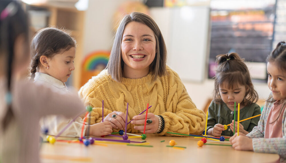 A smiling female educator with a group of children, playing with colourful toys. The teacher is white with short brown hair and wears a yellow knitted jumper.