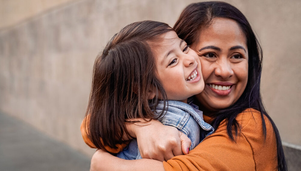 A mum and daughter sharing a happy embrace. Both have dark hair and brown eyes and are looking slightly off camera. The mother is wearing a orange top and the daughter is wearing a denim jacket.