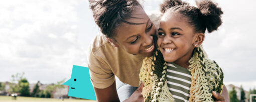 A black mother and her young daughter stand in a field. Goal posts can be seen in the background. The mother has dark hair which is pulled back into a bun and she is bent down hugging her daughter from behind. Her daughter has dark brown hair pulled into two pig tails and wears a green crocheted cardigan. Both are smiling.