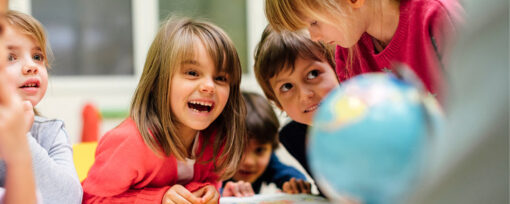 A group of children excitedly gather around a table. They are all primary-aged and appear to be listening to someone off camera. There is a globe on the table indicating they are in a classroom although they are not in school uniform.