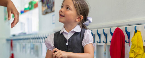 A young white school girl with light brown hair is smiling in the hallway and looking up to talk to someone. She is primary-aged and wearing a white t-shirt with a grey pinafore. Presumably, her school uniform. There is jackets hanging on hooks on the right of the image.