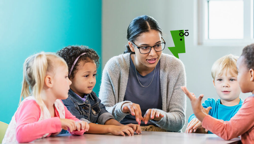 An educator interacting with a group of four children around a table. They are talking and a graphic of a green character shaped like a lightening bolt wearing black glasses can be seen in the background. The character has a shocked expression.