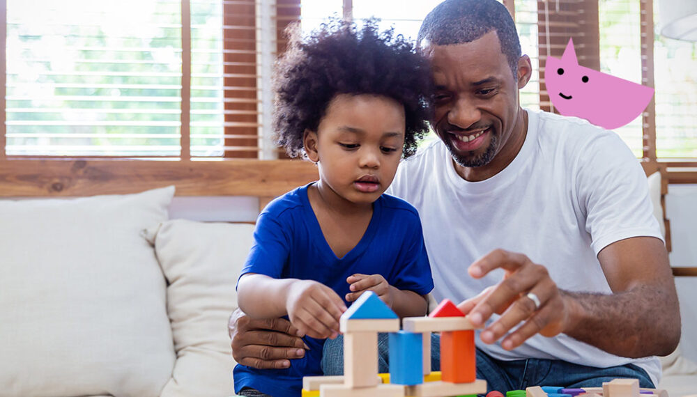 A black man and his son playing with building blocks, with a pink Speech and Language UK character in the background. The son has curly dark hair and wear a blue t-shirt. The father has a buzzcut and wears a white t-shirt. The character is shaped like a pink semi-circle and sits on the right, appearing to be on the fathers shoulder.