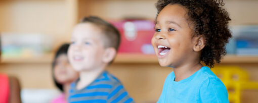 Young black boy sat in classroom smiling. He has curly hair and wears a bright blue t-shirt.