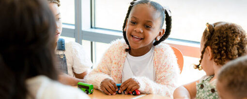 A young black girl playing with a toy train and smiling at a grown-up. The girl has two braids with white bows and a pink fluffy cardigan.