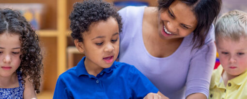 A teacher with three young children. It looks like a nursery and she is showing them something on a page and pointing. It looks like the children and colouring.