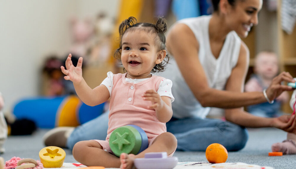 Young child in pink playing at a nursery with blocks. An adult and other children feature playing in the background.