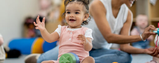 Young child in pink playing at a nursery with blocks. An adult and other children feature playing in the background.